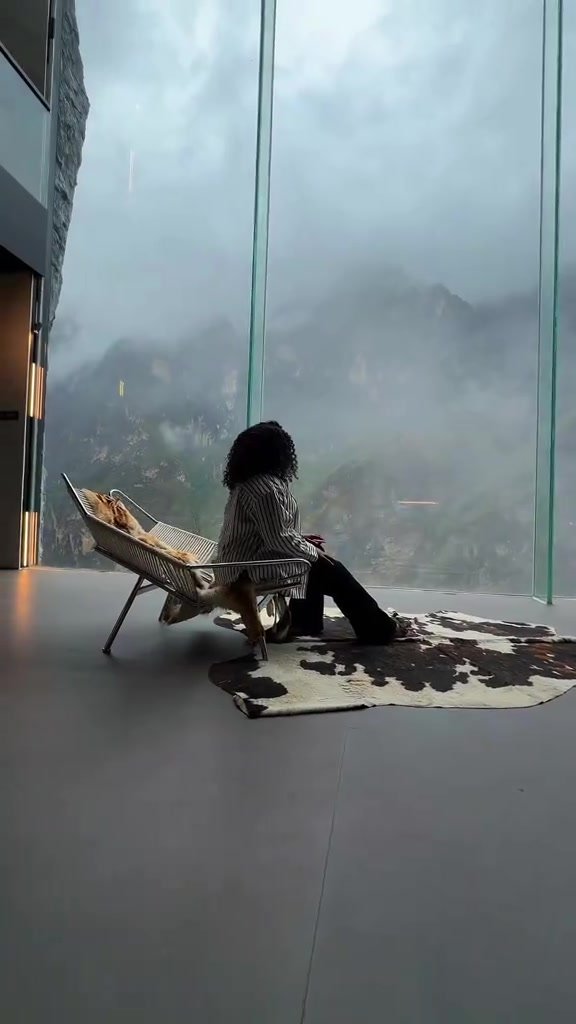 Lounge chair beside panoramic windows and cloud-covered mountains