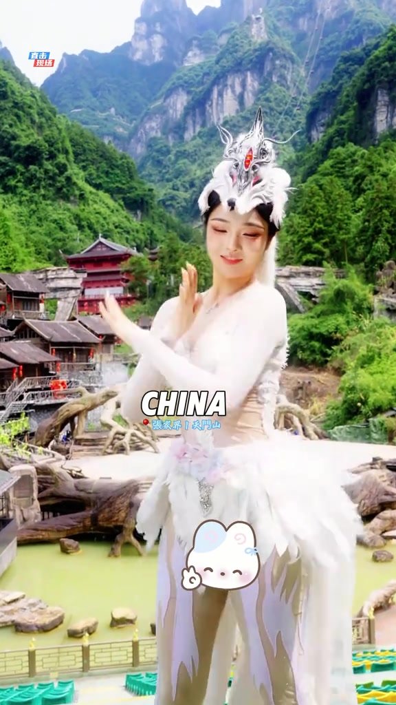 A white-costumed fox-fairy performer dancing before steep green mountains and traditional-style buildings